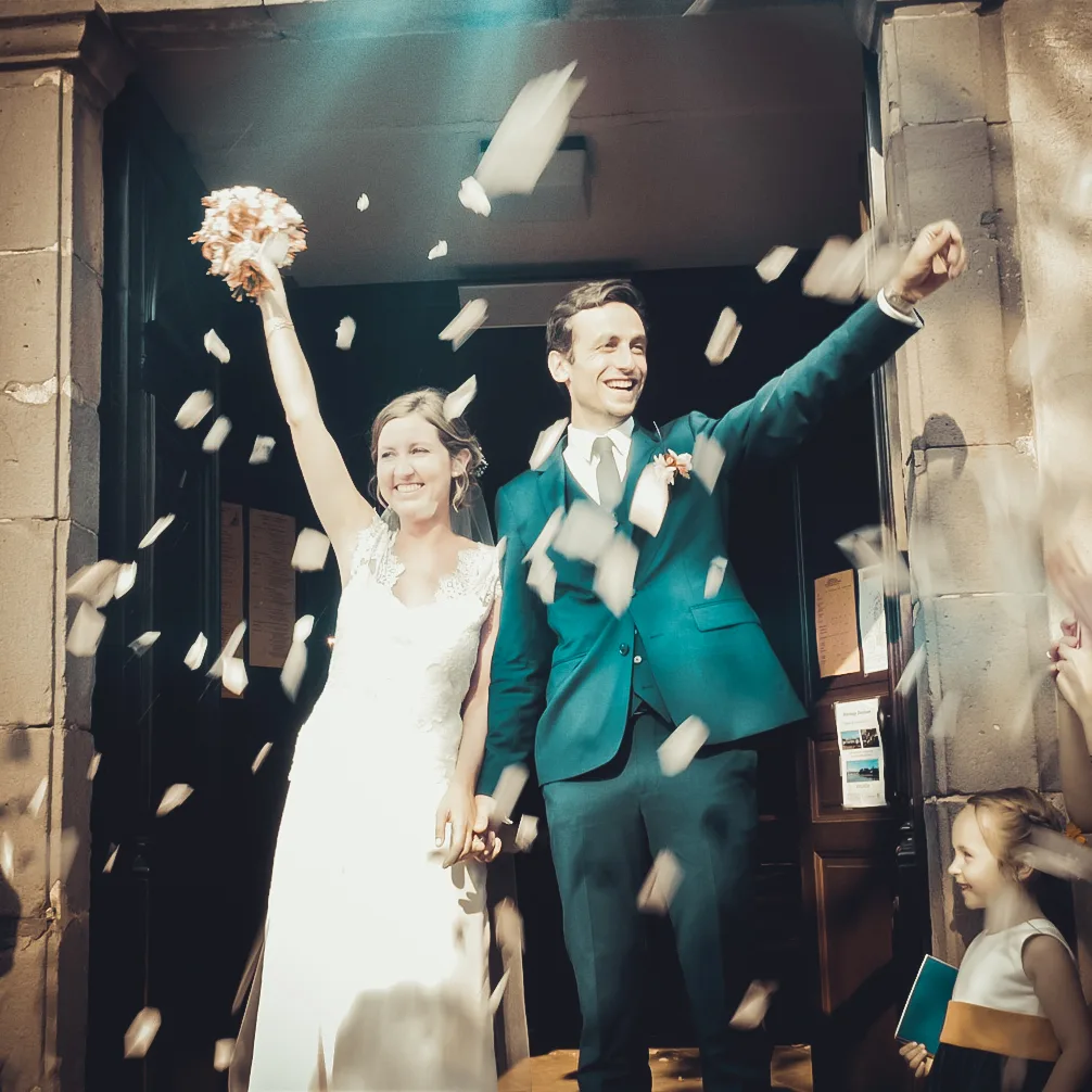 couple de marié sortant de l'église les bras levés sous les pétales de roses.